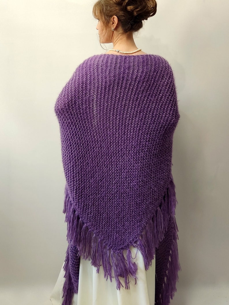 May include: A purple knitted shawl with fringe. The shawl is made of a soft, fuzzy yarn and has a triangular shape.