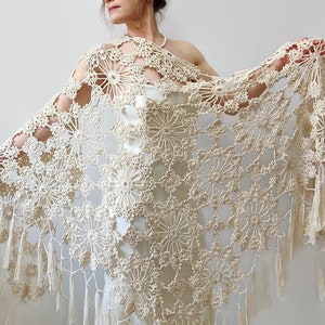 May include: Cream-colored, open-weave crochet shawl with a floral pattern and fringe trim. The shawl is draped over the shoulders, showcasing the intricate design and texture. The delicate, handmade piece is a beautiful accessory.