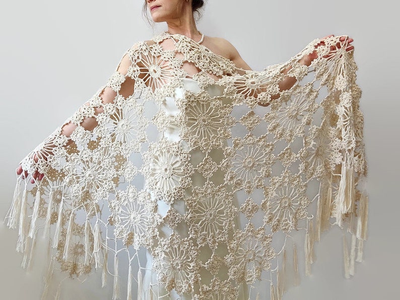 May include: Cream-colored, open-weave crochet shawl with a floral pattern and fringe trim. The shawl is draped over the shoulders, showcasing the intricate design and texture. The delicate, handmade piece is a beautiful accessory.