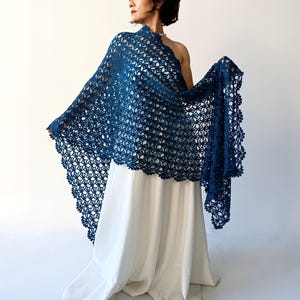 May include: A navy blue crocheted shawl with a floral pattern and scalloped edges. The shawl is draped over a white dress, creating a flowing, elegant look. The openwork design allows the dress to be seen beneath.