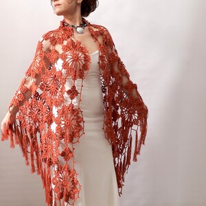 May include: A terracotta crocheted shawl with fringe. The shawl is made with a delicate openwork pattern and has a flowing, elegant drape.