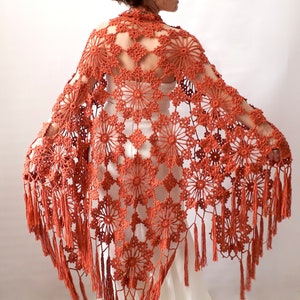 May include: A crocheted shawl with a floral pattern and fringe. The shawl is a burnt orange color.