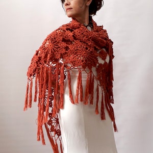 May include: A terracotta crocheted shawl with a floral pattern and fringe. The shawl is draped over a white dress.