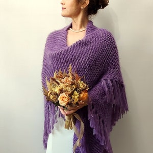 May include: A purple knitted shawl with fringe trim. The shawl is draped over a person's shoulders and they are holding a bouquet of dried flowers.