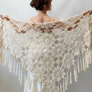 May include: Cream-colored, open-weave crochet shawl with a floral pattern and fringe. The shawl is held open, showcasing the intricate design and texture. The delicate, handmade piece is suitable for special occasions.