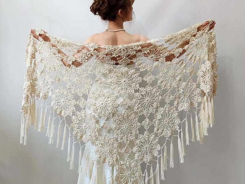 May include: Cream-colored, open-weave crochet shawl with a floral pattern and fringe. The shawl is held open, showcasing the intricate design and texture. The delicate, handmade piece is suitable for special occasions.