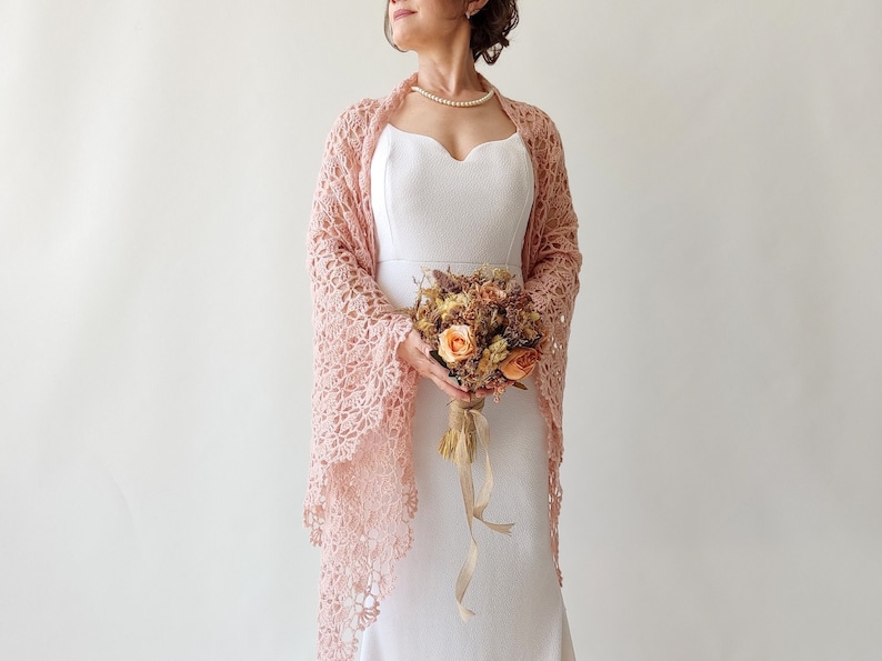 May include: A bride wearing a white dress with a pink crocheted shawl and holding a bouquet of dried flowers.