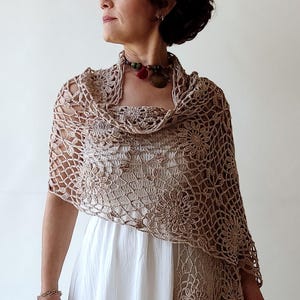 May include: A beige crochet shawl with a delicate floral pattern. The shawl is draped over a person's shoulders and is worn with a white dress.