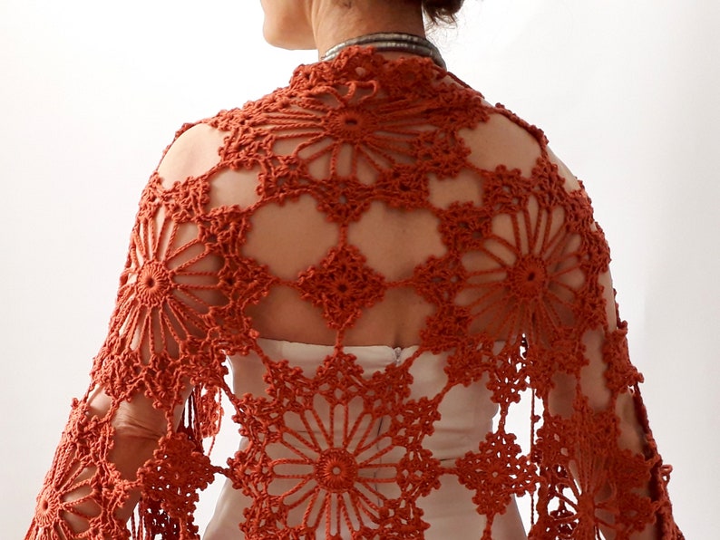 May include: A rust-colored crocheted shawl with a delicate floral pattern. The shawl is made with a lightweight yarn and has a fringed edge.
