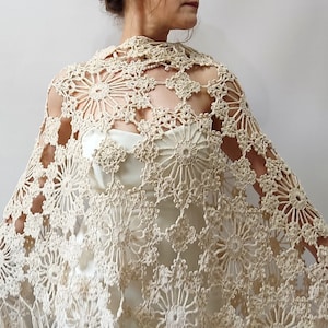 May include: A cream-colored, open-weave crochet shawl with a floral pattern. The shawl is draped over a dress, showcasing the intricate design and texture. The crochet work features circular and geometric shapes, creating a delicate and elegant accessory.