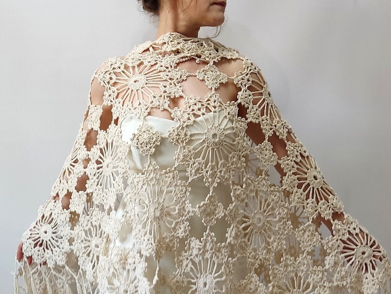 May include: A cream-colored, open-weave crochet shawl with a floral pattern. The shawl is draped over a dress, showcasing the intricate design and texture. The crochet work features circular and geometric shapes, creating a delicate and elegant accessory.