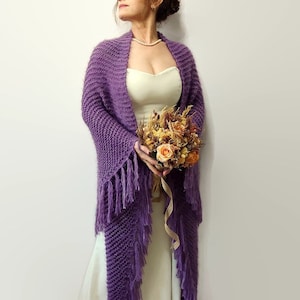 May include: A purple knitted shawl with fringe, worn over a white dress. The shawl is draped over the shoulders and has a loose, flowing fit. The model is holding a bouquet of dried flowers.