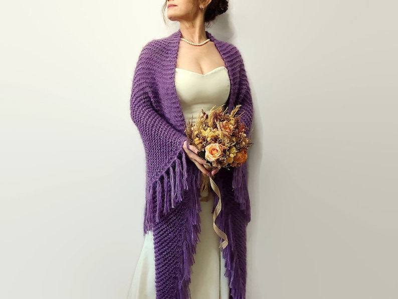 May include: A purple knitted shawl with fringe, worn over a white dress. The shawl is draped over the shoulders and has a loose, flowing fit. The model is holding a bouquet of dried flowers.