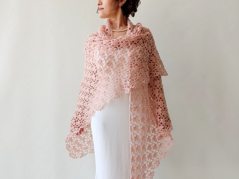 May include: A delicate pink crocheted shawl with a lacy design. The shawl is draped over a person's shoulders and is worn with a white dress.