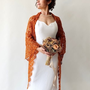 May include: A woman wearing a white dress and a burnt orange crochet shawl holds a bouquet of dried flowers.