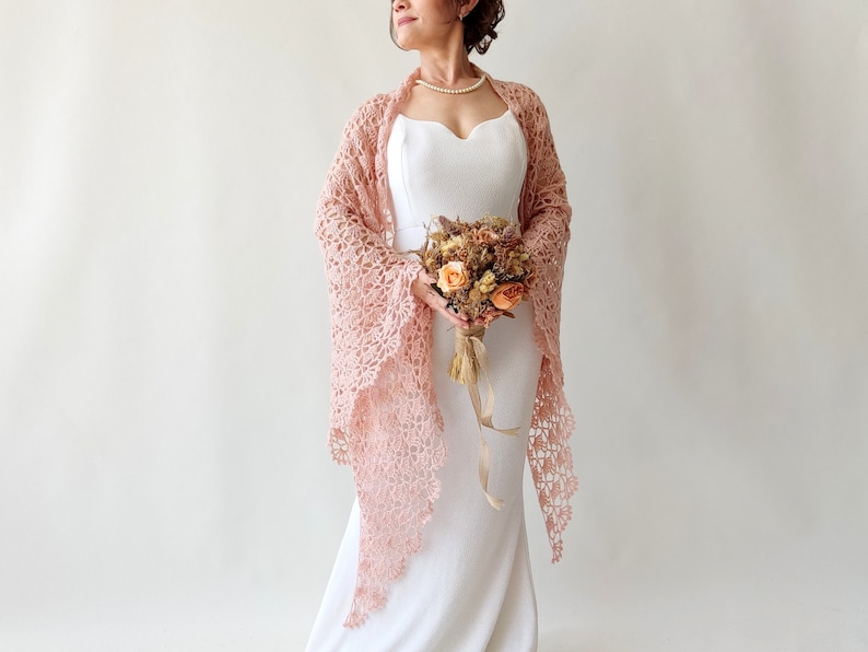 May include: A woman wearing a white dress with a delicate pink crocheted shawl. She is holding a bouquet of dried flowers.