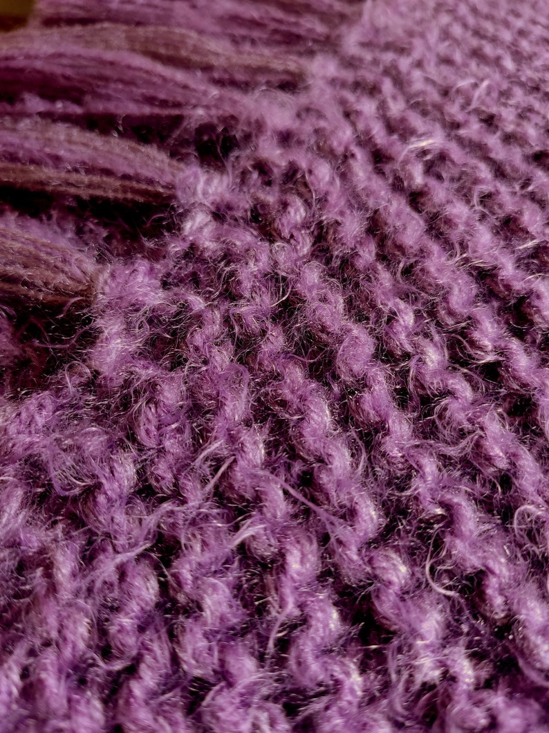 May include: Close-up of a purple fuzzy knitted fabric. The fabric has a soft, fluffy texture and a deep, rich purple color.