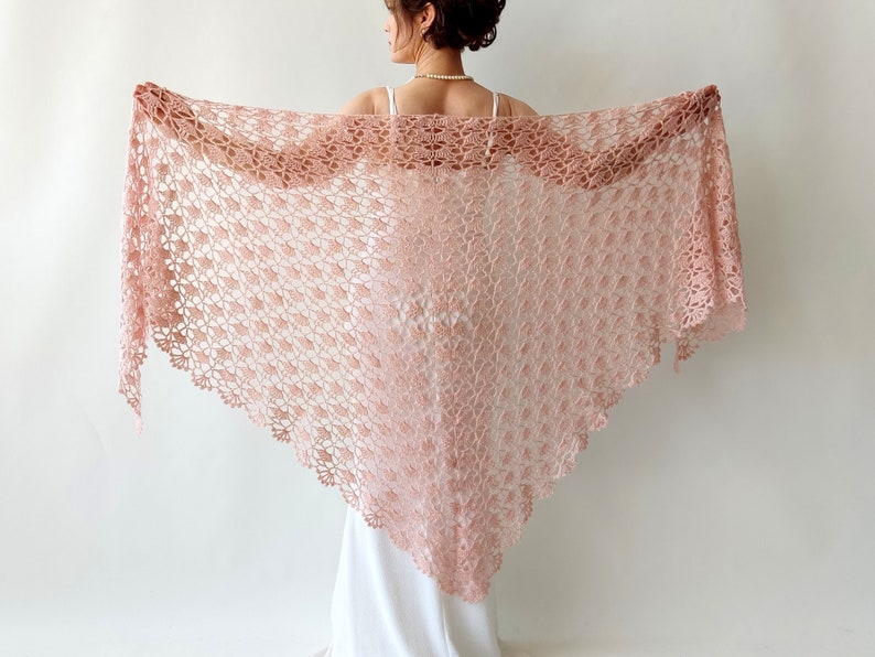 May include: A delicate blush pink crochet shawl with a lacy pattern. The shawl is draped over the shoulders of a person wearing a white dress.