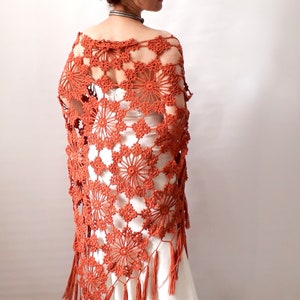 May include: A terracotta crocheted shawl with a floral pattern and fringe. The shawl is draped over a white dress.