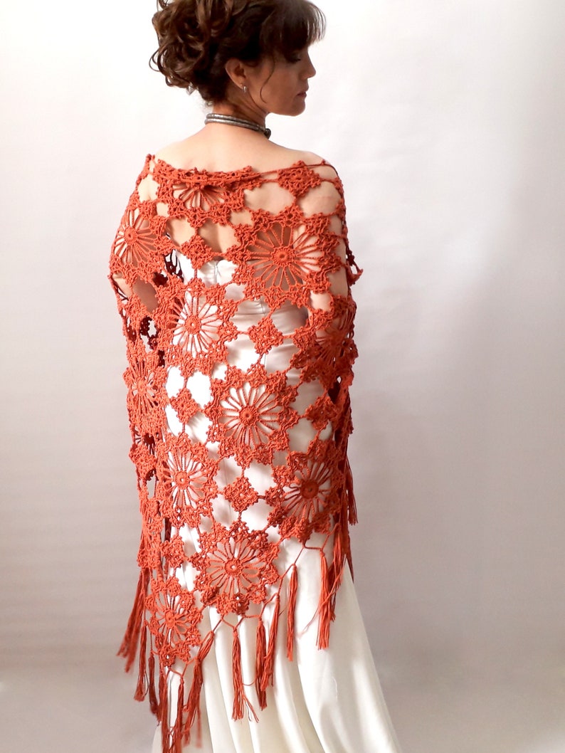 May include: A terracotta crocheted shawl with a floral pattern and fringe. The shawl is draped over a white dress.