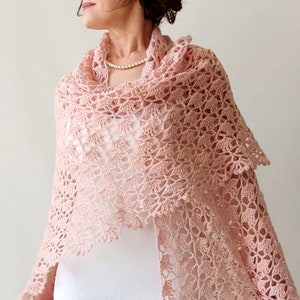 May include: A delicate blush pink crochet shawl with a lacy pattern. The shawl is draped over a person's shoulders and is a beautiful accessory for any occasion.