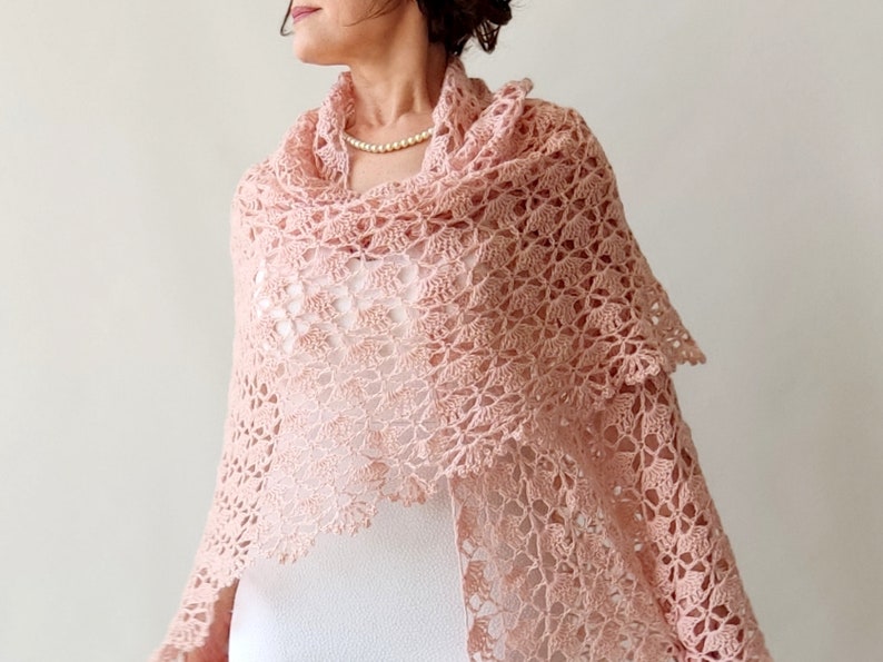 May include: A delicate blush pink crochet shawl with a lacy pattern. The shawl is draped over a person's shoulders and is a beautiful accessory for any occasion.