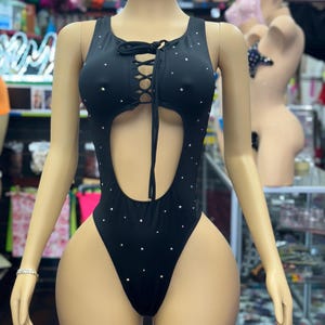 May include: Black one-piece swimsuit with a lace-up front and a large cutout. The swimsuit is embellished with small, sparkling rhinestones. The mannequin is wearing a bracelet on its left wrist. Other lingerie items are visible in the background.