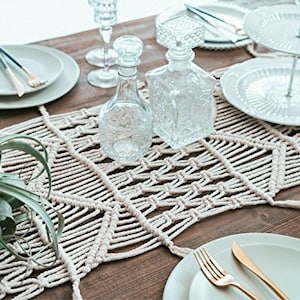 May include: A white macrame table runner with a geometric pattern. The runner is laid on a wooden table with white plates, glasses, and a plant.