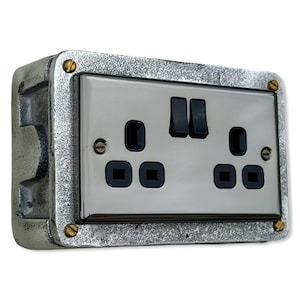 Puede incluir: Un enchufe eléctrico de color plateado con dos enchufes y dos interruptores. El enchufe está montado en un marco de metal gris texturizado y asegurado con tornillos de color latón. Los enchufes son negros y cuadrados.