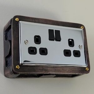May include: A retro-style electrical socket with a polished chrome faceplate. It has two black switches and four black plug sockets. The surrounding frame is a dark, aged bronze colour, with brass-coloured screws and corner accents.