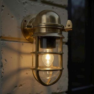 May include: A brass wall-mounted light fitting with a clear glass enclosure and protective cage. The light is illuminated, emitting a warm glow. The fitting is mounted on a white brick wall. The design is reminiscent of nautical or industrial styles.