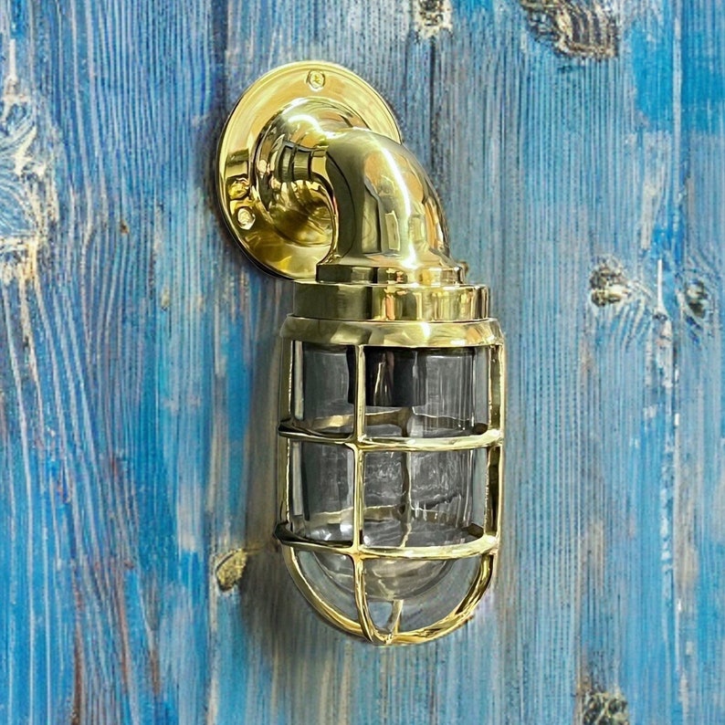 Puede incluir: Aplique de pared de lat&oacute;n de estilo n&aacute;utico con una carcasa de vidrio transparente. La l&aacute;mpara tiene una placa trasera redondeada, un brazo curvo y una estructura en forma de jaula alrededor del vidrio. Montado sobre una superficie de madera azul.