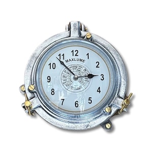 May include: A round, silver-colored clock with a white face and black numerals. The clock has a nautical design, with a circular frame and brass accents. The clock face includes the text "MAXLUME" and a telegraph dial. The clock measures approximately 6 inches in diameter.