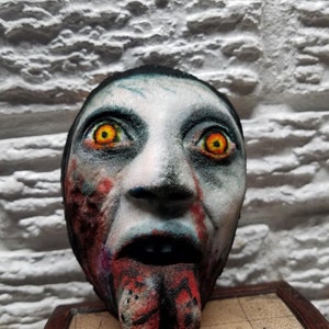 May include: A 3D printed, hand-painted, realistic-looking zombie head with a long, red tongue sticking out. The head has a white, gray, and black painted face with red and blue accents. The eyes are painted yellow with black pupils.