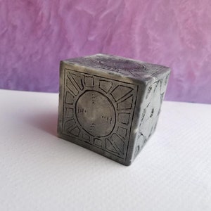 Może przedstawiać: Szara kostka z detalicznym wzorem na każdej ścianie. Wzór przedstawia okrągły wzór z liniami i mniejszym kółkiem w środku.