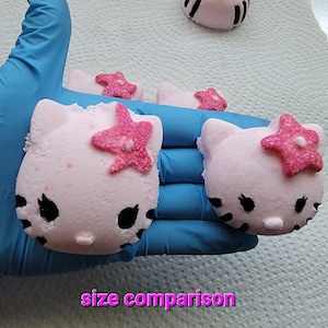 Könnte beinhalten: Größenvergleich von rosa Badebomben in Form einer Comic-Katze mit schwarzen Augen und einer rosa Blume obenauf. Die Badebomben liegen auf einer weißen Oberfläche.
