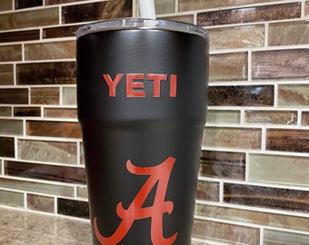 Op de afbeelding: Een zwarte Yeti-beker met een rode "A" en het woord "YETI" in rode letters. Een witte rietje zit in de beker.