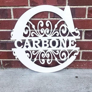 K&ouml;nnte beinhalten: Ein wei&szlig;es Metallschild mit einem dekorativen Schriftrollenmuster und dem Nachnamen "CARBONE" in einem geteilten Monogramm-Stil.