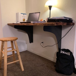 May include: A wooden desk with black metal brackets, a laptop, a lamp, and a backpack on the floor. The desk is mounted to the wall and has a small stool in front of it.