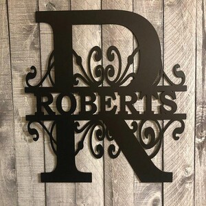 K&ouml;nnte beinhalten: Schwarzes Metallmonogramm mit dem Buchstaben "R" und dem Namen "ROBERTS" in einer dekorativen Schriftart. Der Buchstabe "R" hat ein dekoratives Schn&ouml;rkeldesign.