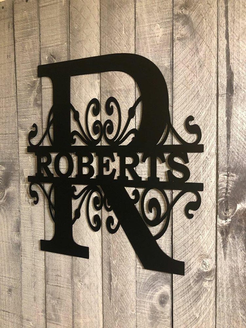 K&ouml;nnte beinhalten: Schwarzes Metall-Monogrammschild mit dem Buchstaben "R" und dem Nachnamen "ROBERTS" in einer dekorativen Schriftart. Der Buchstabe "R" ist mit einem dekorativen Schriftrollenmuster verziert.