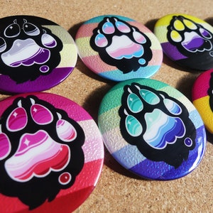 Könnte beinhalten: Satz von zehn 2,5 cm großen Anstecknadeln mit einem Pfotenabdruck in verschiedenen Farben von Pride-Flaggen. Die Flaggen umfassen die Ace-, Aro-, Trans-, Bi-, Lesben-, Pan-, Genderqueer-, Non-Binary- und Genderfluid-Flaggen.