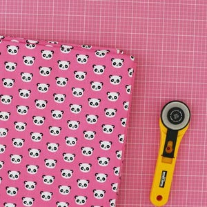 May include: A folded fabric featuring a panda bear pattern in black and white on a pink background. A yellow rotary cutter with a black blade is on the right. The background is a pink cutting mat.