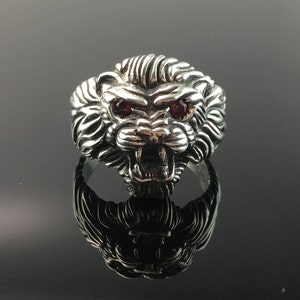 Può includere: Un anello in argento a forma di testa di leone con occhi di pietre preziose rosse. L'anello è dettagliato con pelliccia testurizzata e un'espressione ringhiante.