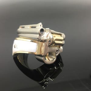 May include: A gold and silver ring with a detailed design of a gun. The ring has a white inlay on the band.