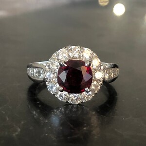 May include: A silver ring with a large, round, deep red gemstone at its centre, encircled by a halo of smaller, clear gemstones. The ring's band has additional clear gemstones on each side, creating a sparkling, elegant design.