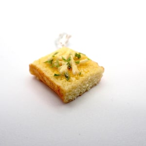 Könnte beinhalten: Ein kleiner, quadratischer Anhänger, der einem Stück Toastbrot ähnelt, mit geschmolzenem Käse und grünen Kräutern belegt. Das Brot hat eine goldbraune Kruste. Der Anhänger ist wahrscheinlich für eine Halskette, die Maße sind in Zentimetern.