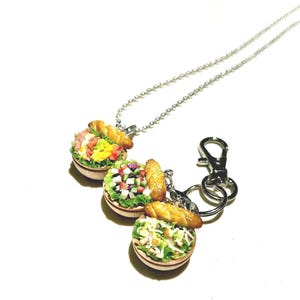 May include: Miniature salad-themed jewelry and keychain set. The set includes three small, detailed salad bowls with various toppings, each topped with a miniature baguette. One bowl is on a silver chain necklace, and another is attached to a keychain.