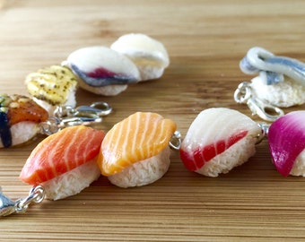May include: A collection of sushi-themed keychains. Each keychain features a miniature sushi piece, including salmon, tuna, and other varieties, atop a bed of rice. The sushi pieces have realistic details and are attached to silver-colored clasps.