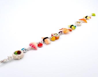 May include: A silver chain bracelet featuring colorful sushi charms. The charms include various sushi rolls, nigiri, and other Japanese food items. The bracelet is on a white background.